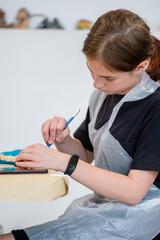 Young artist focused on painting a decorative object with precision. Wearing a protective apron,...