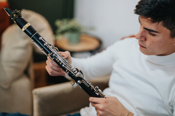 A neatly dressed young man closely examines the intricate details of his clarinet in a warm,...