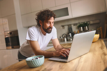 A man with curly hair is focused on his laptop placed on a wooden kitchen table. He snacks on a...