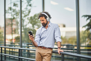 A businessman in a light blue shirt and tan pants stands by a railing, enjoying music on headphones...