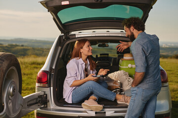 Two friends relax in a green field, sharing snacks and drinks from the back of an SUV while enjoying their time outdoors in nature under clear skies.