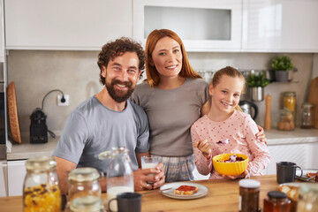 A joyful family shares breakfast in a warm kitchen, filled with smiles and laughter. The daughter enjoys her bowl of yogurt, while the parents toast with drinks.