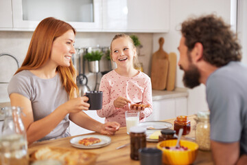 A family gathers around the kitchen table, sharing a joyful breakfast. They smile, drink coffee and milk, and savor pastries while enjoying each other's company on a relaxed morning.