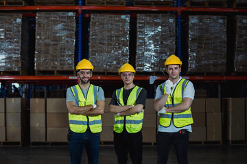 Three confident male warehouse workers in safety vests and helmets stand with arms crossed in front of stacked storage shelves under factory lighting.