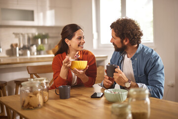 A couple shares a joyful moment in their home office, enjoying breakfast and coffee while seated at a wooden table surrounded by a warm atmosphere and kitchen decor.