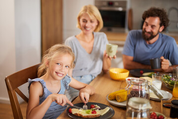 A family engages in a cheerful breakfast, with a girl smiling and eating, while her parents watch with happiness in a warm kitchen atmosphere.