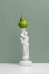 Green pear with pins inserted mounted on white female statue pedestal over muted green wall. Concept of pop art, visual metaphor, health discomfort, art parody, food and symbolism.