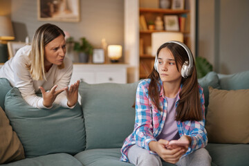 A teenager sits on a couch wearing headphones, showing disinterest as a parent gestures...