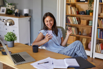 A woman sits comfortably in a bright home office, smiling at her smartphone while holding reading...