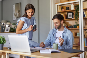 Two individuals engage in a productive work session in a home office. One person sits at a desk with a laptop, while the other stands beside holding a coffee mug.