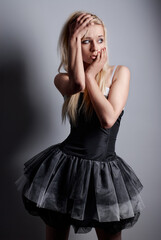 Fototapeta premium Bblonde woman wearing stylish black dress poses in studio, showcasing emotion and intense expressions of shock or worry. Muted backdrop highlights the contrast with the elegant garment.
