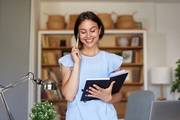 A woman enjoys a productive moment in her home office, holding a notebook and a pen, smiling as she...