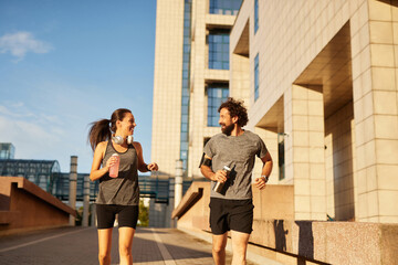A man and woman are jogging together on a city path lined with modern buildings. They are smiling,...
