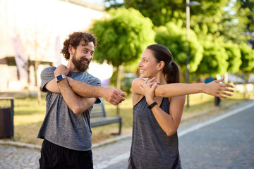 Two individuals engage in stretching exercises on a sunny day in a park, preparing for a run while...