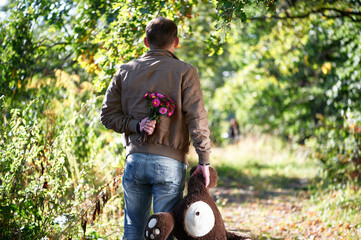 Man holding a bouquet of flowers and a teddy bear, waiting for his date in a sunlit park path.