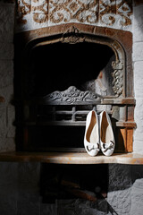 A pair of elegant white shoes with bows is showcased on the hearth of a vintage, ornate fireplace,...