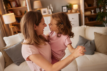 A mother holds her daughter in a warm embrace, both smiling and enjoying a playful dance in a cozy...