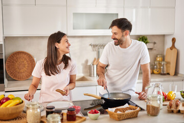 A couple is joyfully engaged in cooking in a well-lit kitchen. One partner stirs a dish on the...