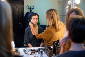 A makeup artist applies products to a seated woman's face in a studio, surrounded by various makeup...