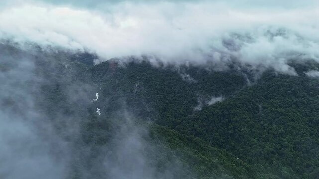 An epic drone view of a cascading waterfall lush mountains and forests topped with misty clouds