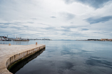 Fototapeta premium A serene waterfront scene featuring calm waters reflecting a cloudy sky. The image captures a stone pier extending into the water, with boats visible in the distance.