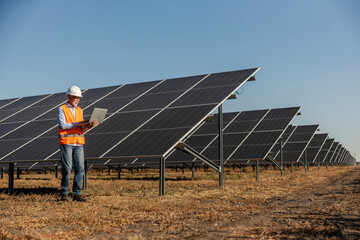 Full length of an old technician using laptop near solar panels at solar plant.