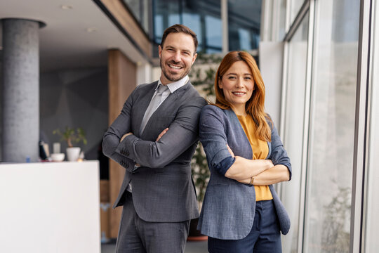 Portrait of businesspeople standing back to back with arms crossed and smiling at camera at corporate office.