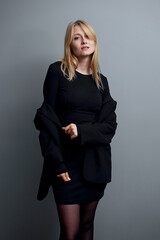 A blonde woman wearing an elegant black ensemble poses confidently against a muted gray background.