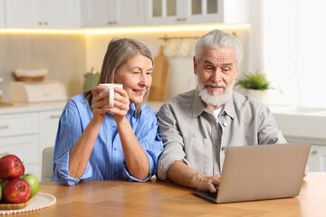 Fototapeta premium Cute elderly couple with laptop at table in kitchen
