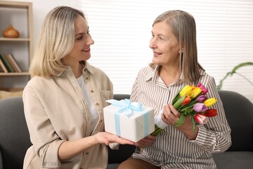 Smiling daughter congratulating her mom with bouquet of tulips and gift on sofa at home. Happy...