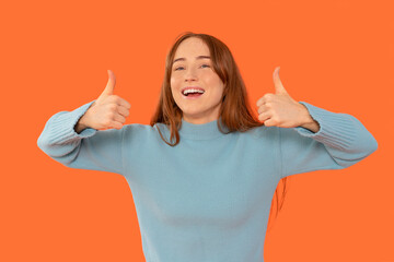 Cheerful young woman giving thumbs up against bright orange background, showcasing enthusiasm and...