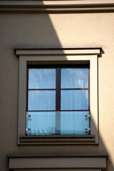 A close-up of a modern window on a beige building wall, partially covered by a diagonal shadow. The window has dark brown wooden frames with four glass panes and a small glass balcony guard. The refle