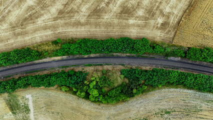 Aerial view of a single railway track cutting through a lush landscape, bordered by dense green trees on both sides. The track is laid on gravel and curves gently across the terrain, with brownish gra © VladyslavShcherbakov