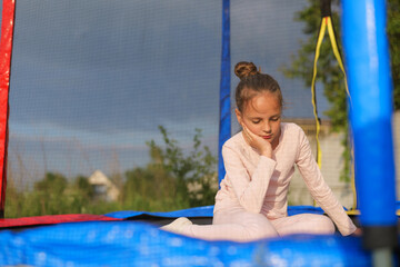 A young girl with her hair in a bun sits thoughtfully on a trampoline, resting her chin on her...