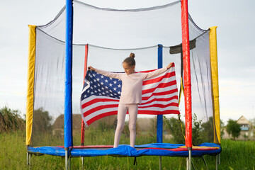 A young girl stands proudly on a trampoline, holding an American flag with outstretched arms, set...