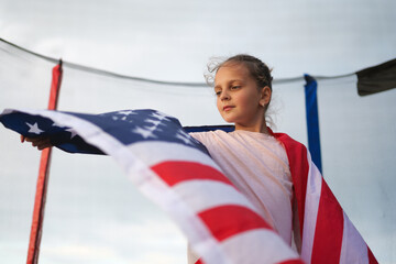 A young girl with a thoughtful expression, draped in an American flag, stands on a trampoline with...
