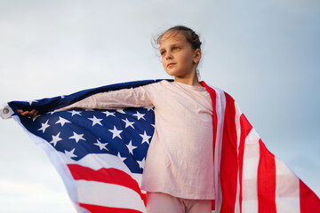 A young girl with a determined and thoughtful expression, wrapped in an American flag, looks into...