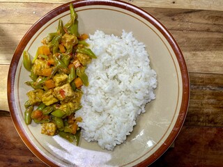 a plate on a wooden table with full of white rice and a side dish of stir-fried stringbean vegetables and tempeh ready to eat, home cooking that is budget-friendly and easy to cook, a breakfast menu