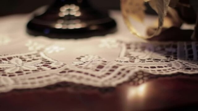 Close-up of a lace doily on a wooden surface, with a blurred lamp base and floral element in the background.