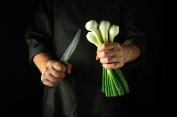 A chef stands ready to chop green onions, holding a bunch in one hand and a sharp knife in the...