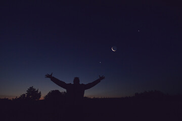Silhouette of a man and countryside under the Moon, stars and planets in blue hour time.