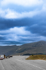 Winding mountain road passes parked vehicles and people under a brooding, cloud-filled sky. Rocky...