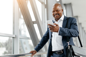 Smiling black businessman checking smartphone while leaning against airport railing. Relaxed and stylish, he holds briefcase and prepares for a professional journey ahead, copy space