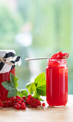 Fresh homemade red currant jam in jar with currants and green leaves on wooden table against light bokeh background. Vertical close-up with space for text.