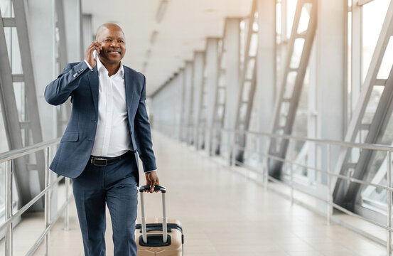 Black business traveler talking on phone while walking with suitcase at airport. Wearing formal suit and bright expression, engaged in smooth departure, copy space