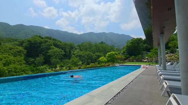 A fit man in his 30s enjoys swimming alone in a scenic infinity pool at a Gapyeong resort, surrounded by lush greenery and mountains, capturing the essence of summer relaxation.