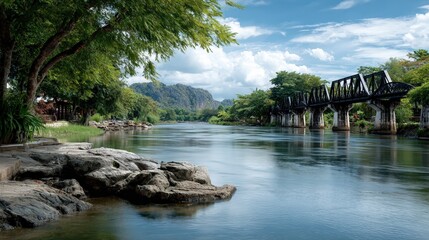 A tranquil river flows past an imposing railway bridge Lush green trees line the rocky bank with distant mountains and a