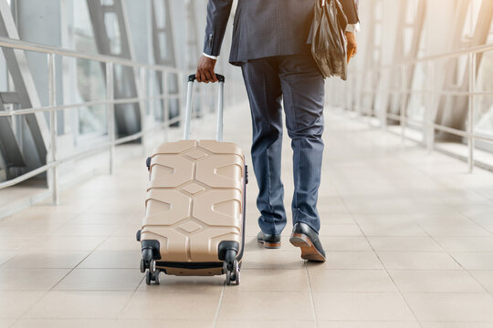 Cropped shot of businessman walking in airport with beige suitcase. Confident steps, smart suit, and briefcase signal his executive presence and readiness to travel, copy space