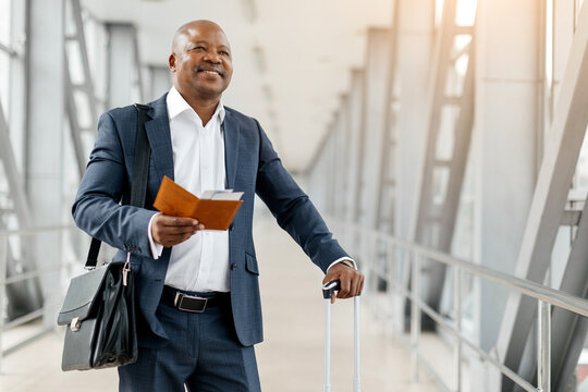 Confident black businessman holding passport and smiling while standing in airport terminal. African American male ready for takeoff, with calm demeanor and suitcase by his side