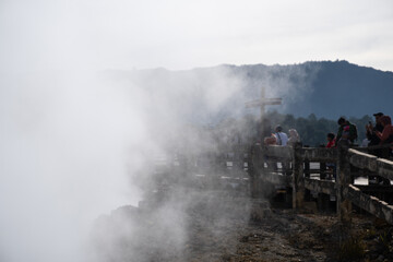 Fototapeta premium Banjarnegara, Indonesia-July 31, 2022: Tourists on wooden pathways observing the steaming Kawah Sikidang volcanic crater in Dieng, surrounded by rugged terrain and green trees under a blue sky.